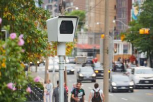 Editorial illustration of a Toronto neighbourhood street with a visible security camera, showing a portrayal of how residential surveillance could appear in the city.