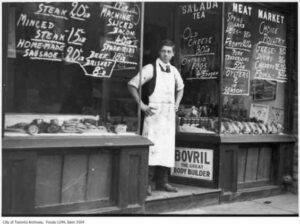 Archival black-and-white photo of Frank James standing in the doorway of a Toronto butcher shop in 1921, with hand-painted meat prices and storefront signs in the window.